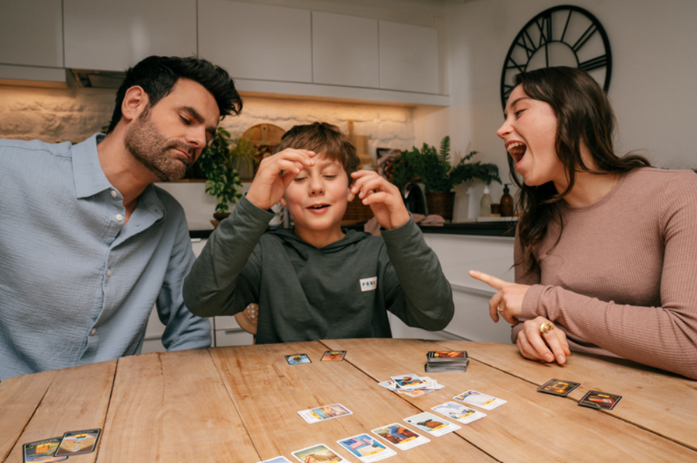 Famille de 3 personnes (deux adultes et un enfant) jouant au jeu de société timeline assis autour d'une table en bois