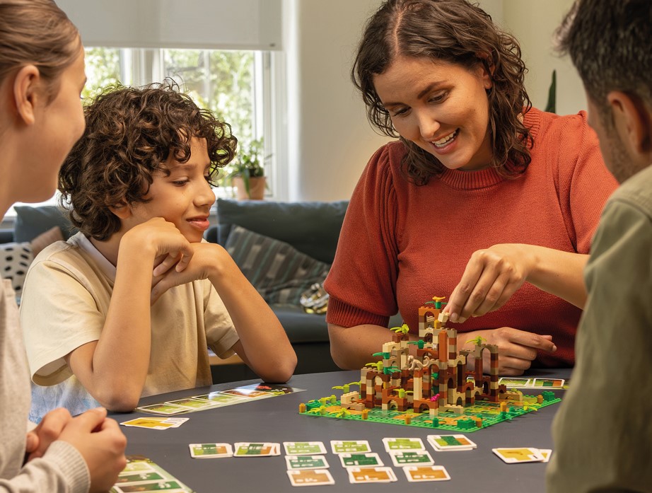 3 adultes et un enfant jouent en famille au jeu de société Monkey Palace avec une structure en briques Lego au milieu de la table.