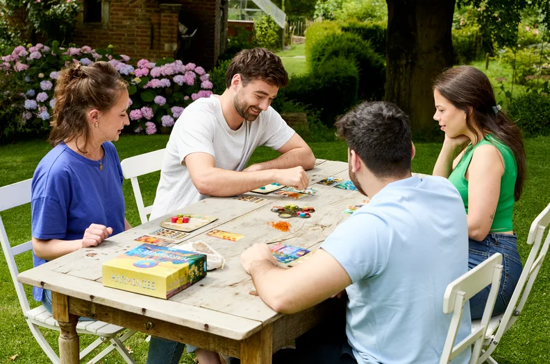 4 personnes jouent à un jeu de société en extérieur dans un jardin sur une table en bois.